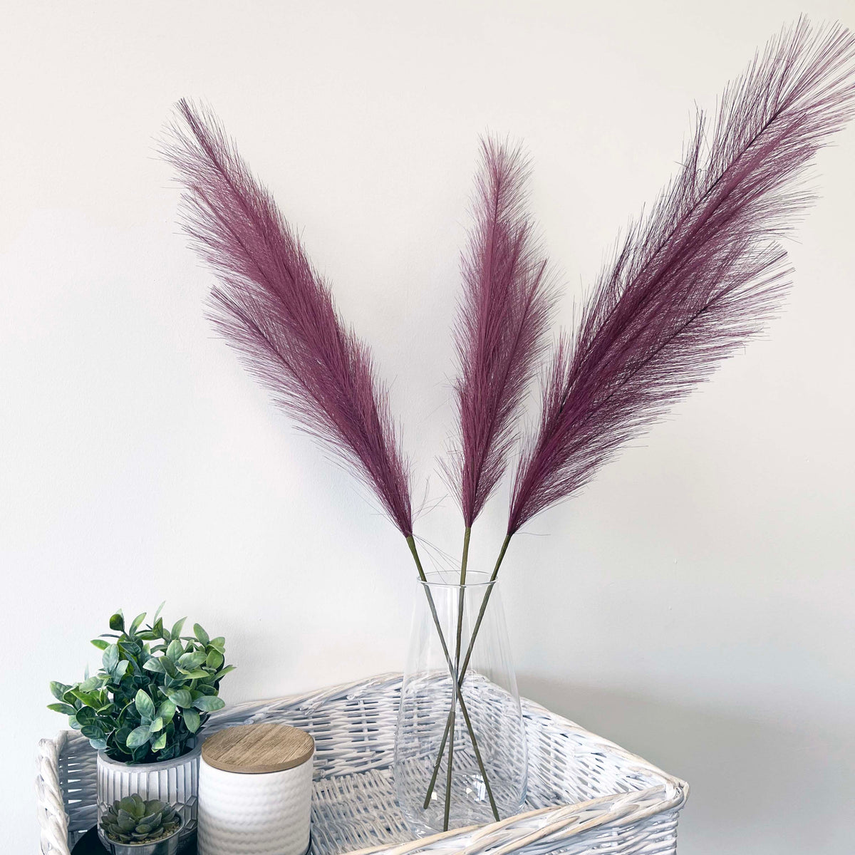 Scarlet Red Faux Pampas Grass Stem in a glass vase, sitting in a wicker table, on a white background.