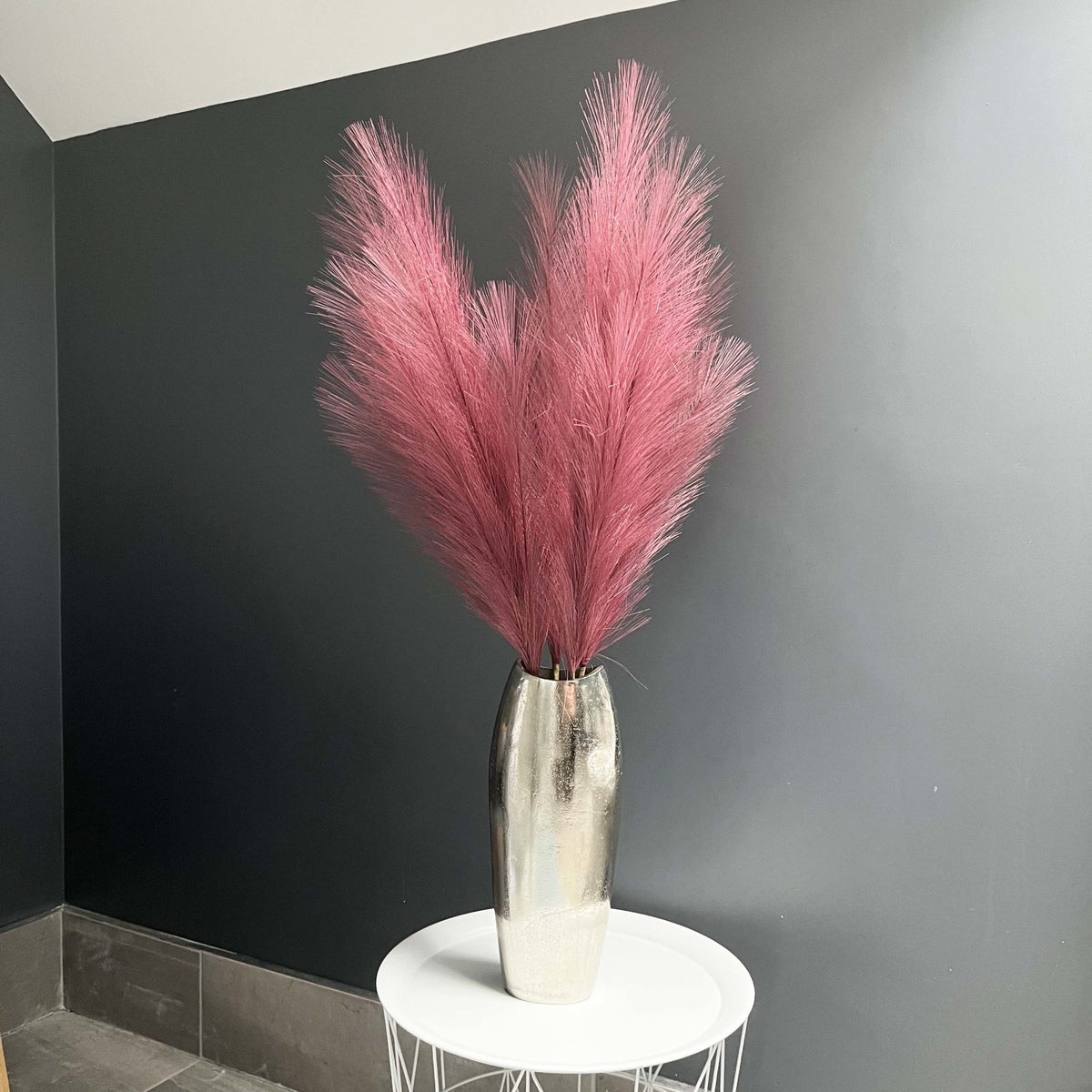 Scarlet Red Faux Pampas Grass Stem in a chrome vase with a dark grey background, on a white table.
