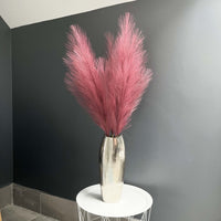 Scarlet Red Faux Pampas Grass Stem in a chrome vase with a dark grey background, on a white table.
