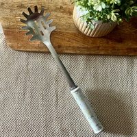 A metal spaghetti server with a white floral-patterned handle rests diagonally on a textured beige and gray herringbone tablecloth, positioned in front of a wooden cutting board with a potted green plant in the background.