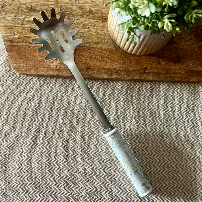 A metal spaghetti server with a white floral-patterned handle rests diagonally on a textured beige and gray herringbone tablecloth, positioned in front of a wooden cutting board with a potted green plant in the background.