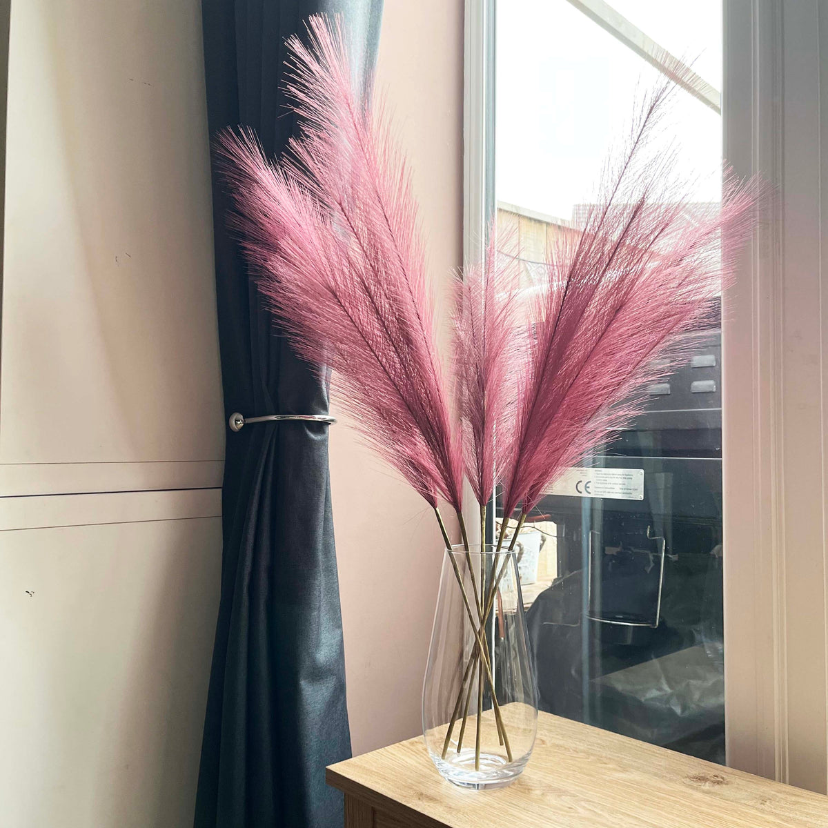 Scarlet Red Faux Pampas Grass Stem in a glass vase on a table next to window.