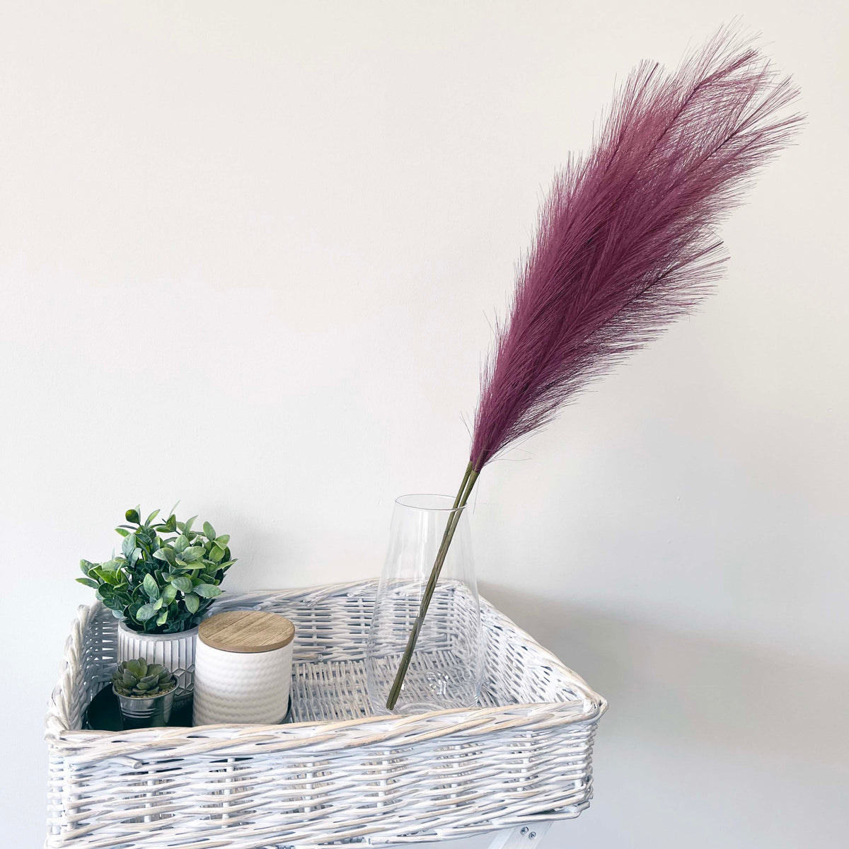 Scarlet Red Faux Pampas Grass Stem in a glass vase, sitting in a wicker table,  on a white background. 