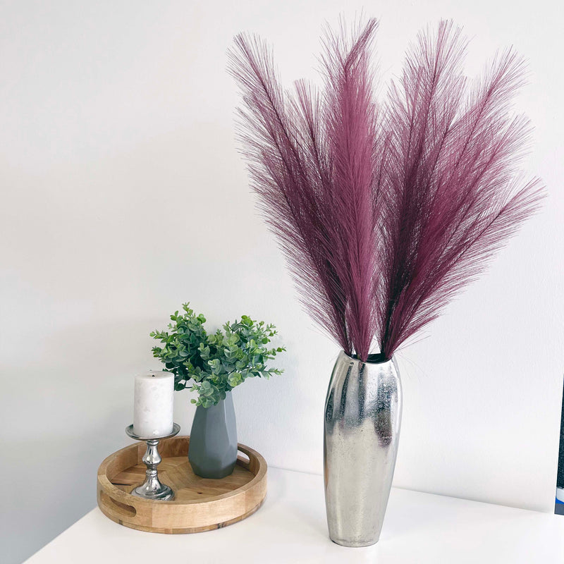Scarlet Red Faux Pampas Grass Stem in a chrome vase with a white background, on a white table. With a wooden tray with plant and candle.