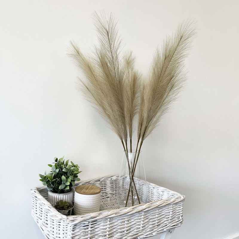 Bleached Faux Pampas Grass Stems in glass vase on white wicker table, with green plant and candle.