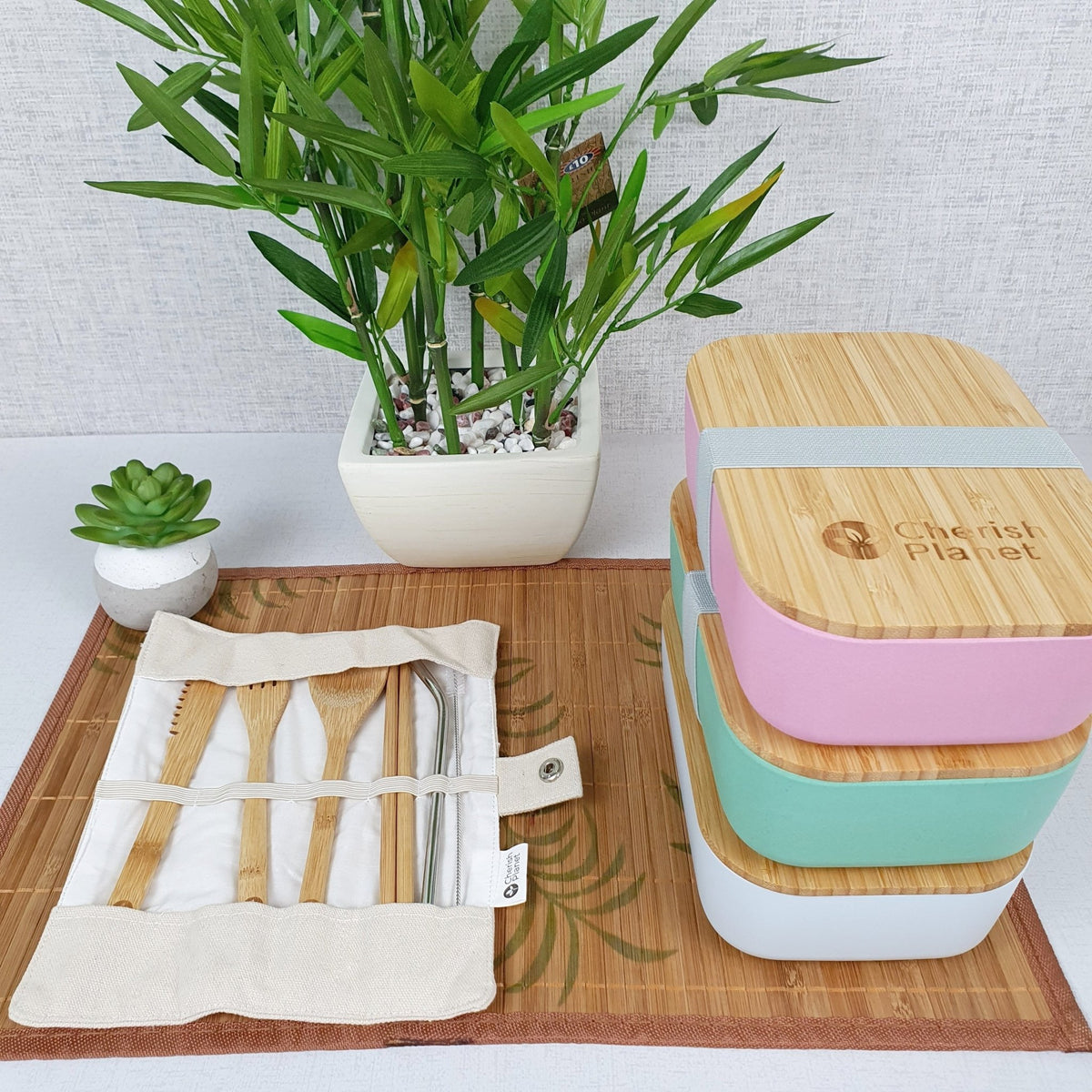 White, green and pink Eco Bamboo & Wheat Fibre Lunch / Bento Box, stacked on top of each other, and cutlery set on white background, with green plant