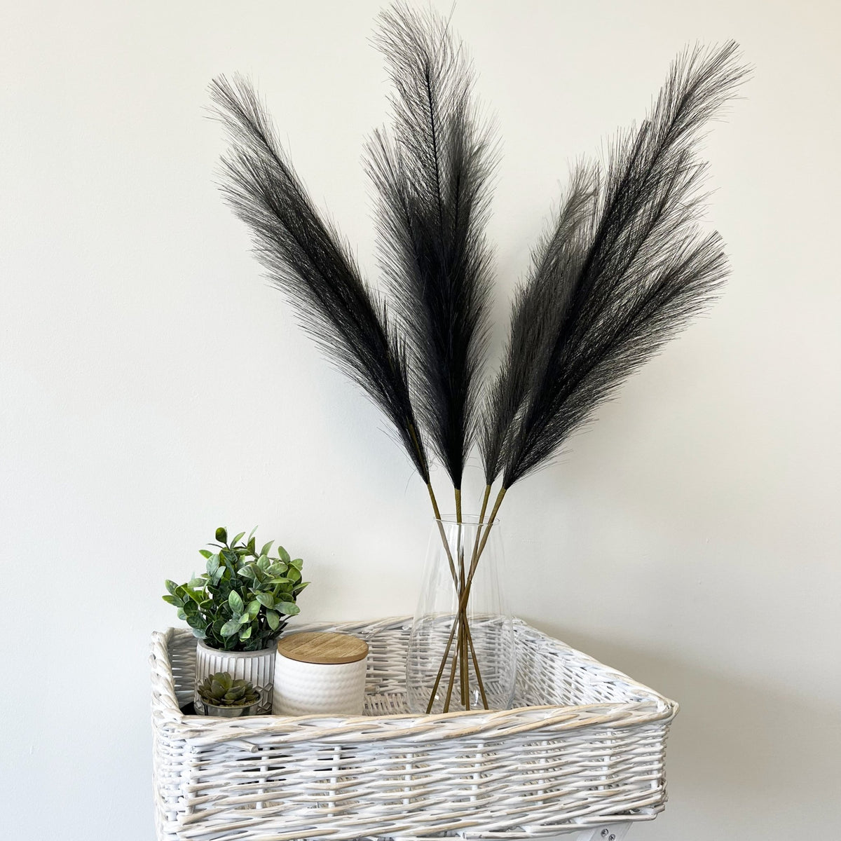 Grey Faux Pampas Grass Stems in glass vase on wicker table with plant and candle.