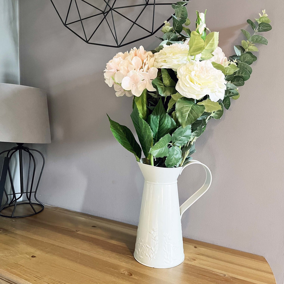White Garden Rose Spray in a water jug vase on a wooden table with black geometric mirror and lamp
